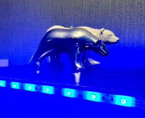 A large bright white Polar Bear Tea Light Holder that has a lit bright white Tea Light coming out of the top of it next to a medium silver Polar Bear behind a small cylindrical frosted glass jar full of some bright blue wax, on a black wooden cabinet.