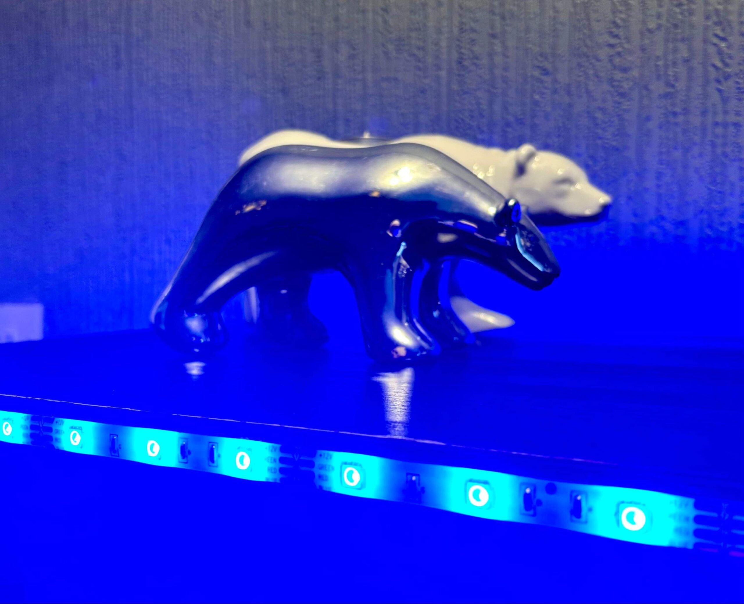 A large bright white Polar Bear Tea Light Holder that has a lit bright white Tea Light coming out of the top of it next to a medium silver Polar Bear behind a small cylindrical frosted glass jar full of some bright blue wax, on a black wooden cabinet.