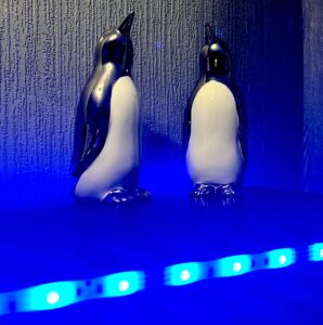 Two tall silver ceramic Penguin ornaments that both have white bellies behind a small cylindrical frosted glass jar full of some bright blue wax, on a black wooden cabinet.