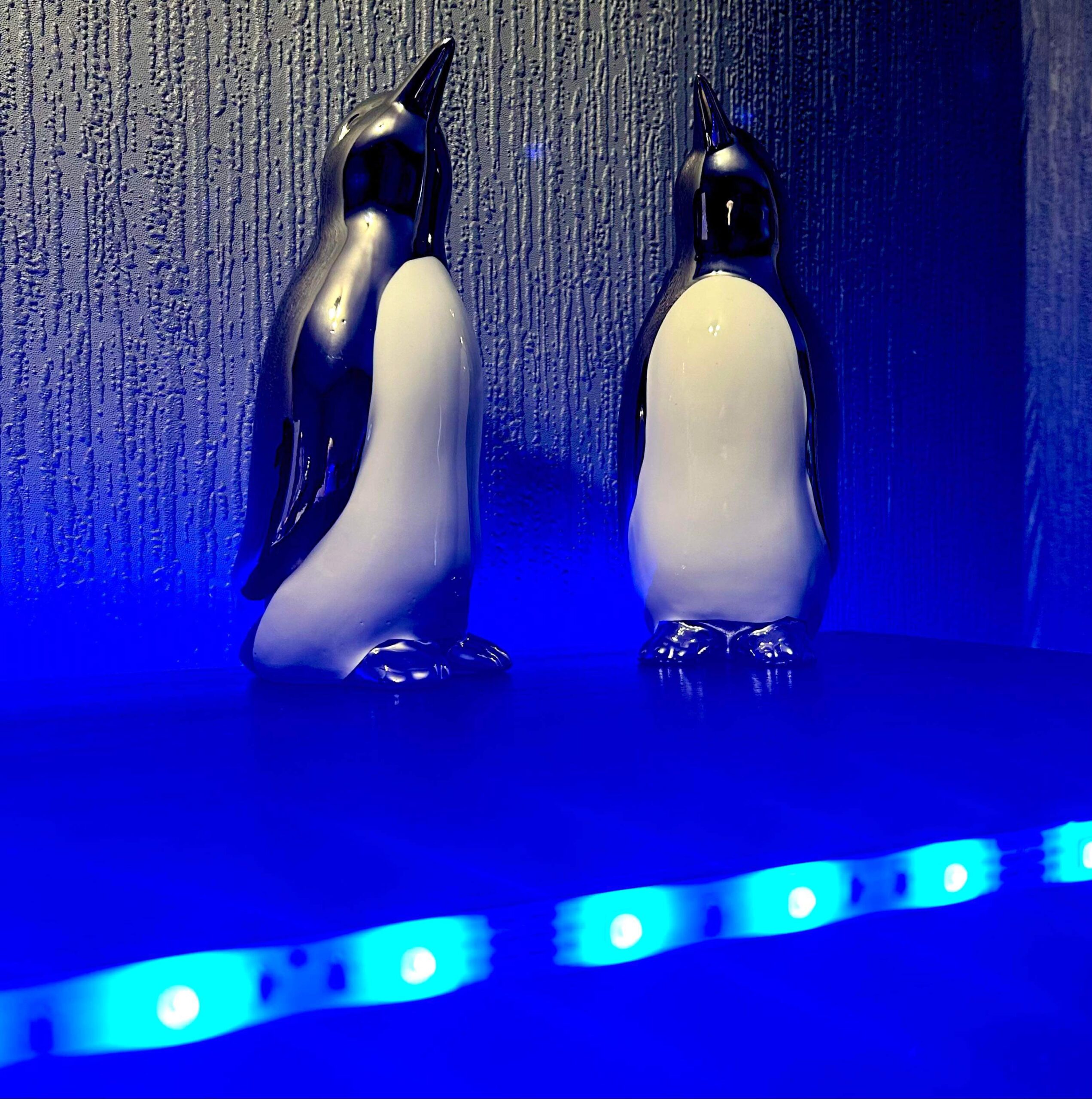 Luke Loves Christmas 2023 | How I've Decorated My Room This Year Two tall silver ceramic Penguin ornaments that both have white bellies behind a small cylindrical frosted glass jar full of some bright blue wax, on a black wooden cabinet.