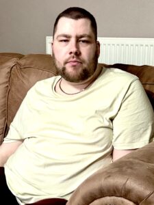 A tall bearded man wearing a bright yellow crew neck t-shirt sat on a dark brown leather sofa.