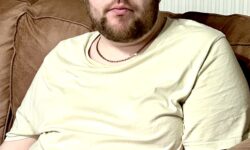 A tall bearded man wearing a bright yellow crew neck t-shirt sat on a dark brown leather sofa.