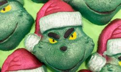 A pile of some medium dark green grinch face shaped bath bombs that all have some bright red Santa’s hats with a white trim and some smirks engraved on them, on a white background.