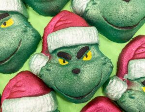 A pile of some medium dark green grinch face shaped bath bombs that all have some bright red Santa’s hats with a white trim and some smirks engraved on them, on a white background.