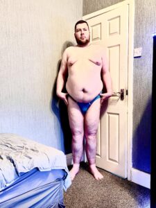 A tall bearded man wearing a pair of bright turquoise blue briefs with a white waistband that has Molly & Tommy written in bold black writing on it stood against a white wooden door.