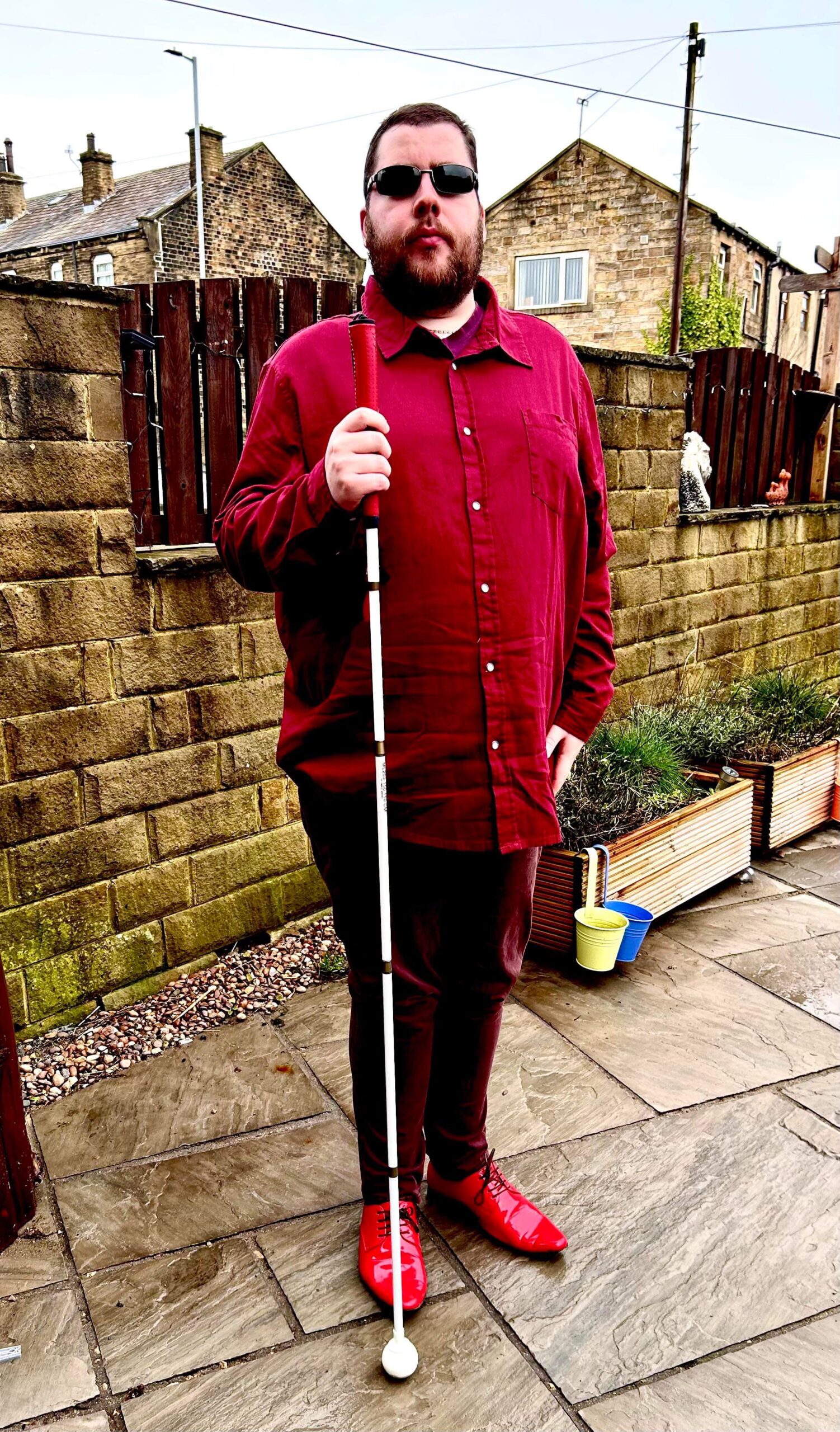 Five Things I Would Like To Achieve In 2025 & Did I Achieve 2024's Goals? A tall bearded man wearing a pair of rectangular silver metal sunglasses with some dark brown lenses, a dark red long sleeve twill shirt, a dark red v-neck t-shirt, a pair of dark red chino trousers and a pair of shiny red shoes holding a bright white cane with a red leather handle stood in front of a dark wooden fence.