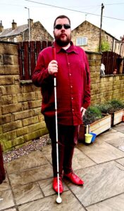 A tall bearded man wearing a pair of rectangular silver metal sunglasses with some dark brown lenses, a dark red long sleeve twill shirt, a dark red v-neck t-shirt, a pair of dark red chino trousers and a pair of shiny red shoes holding a bright white cane with a red leather handle stood in front of a dark wooden fence.