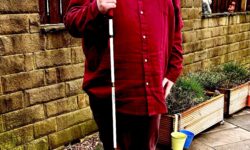 A tall bearded man wearing a pair of rectangular silver metal sunglasses with some dark brown lenses, a dark red long sleeve twill shirt, a dark red v-neck t-shirt, a pair of dark red chino trousers and a pair of shiny red shoes holding a bright white cane with a red leather handle stood in front of a dark wooden fence.