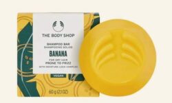 A medium circular bright yellow shampoo bar next to a small square white cardboard box that has The Body Shop Banana Truly Nourishing Shampoo Bar written in dark green writing on it, on a white background.