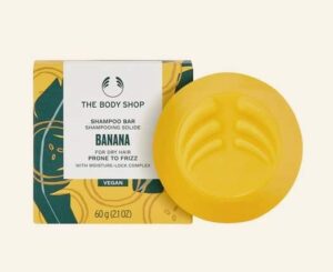 A medium circular bright yellow shampoo bar next to a small square white cardboard box that has The Body Shop Banana Truly Nourishing Shampoo Bar written in dark green writing on it, on a white background.