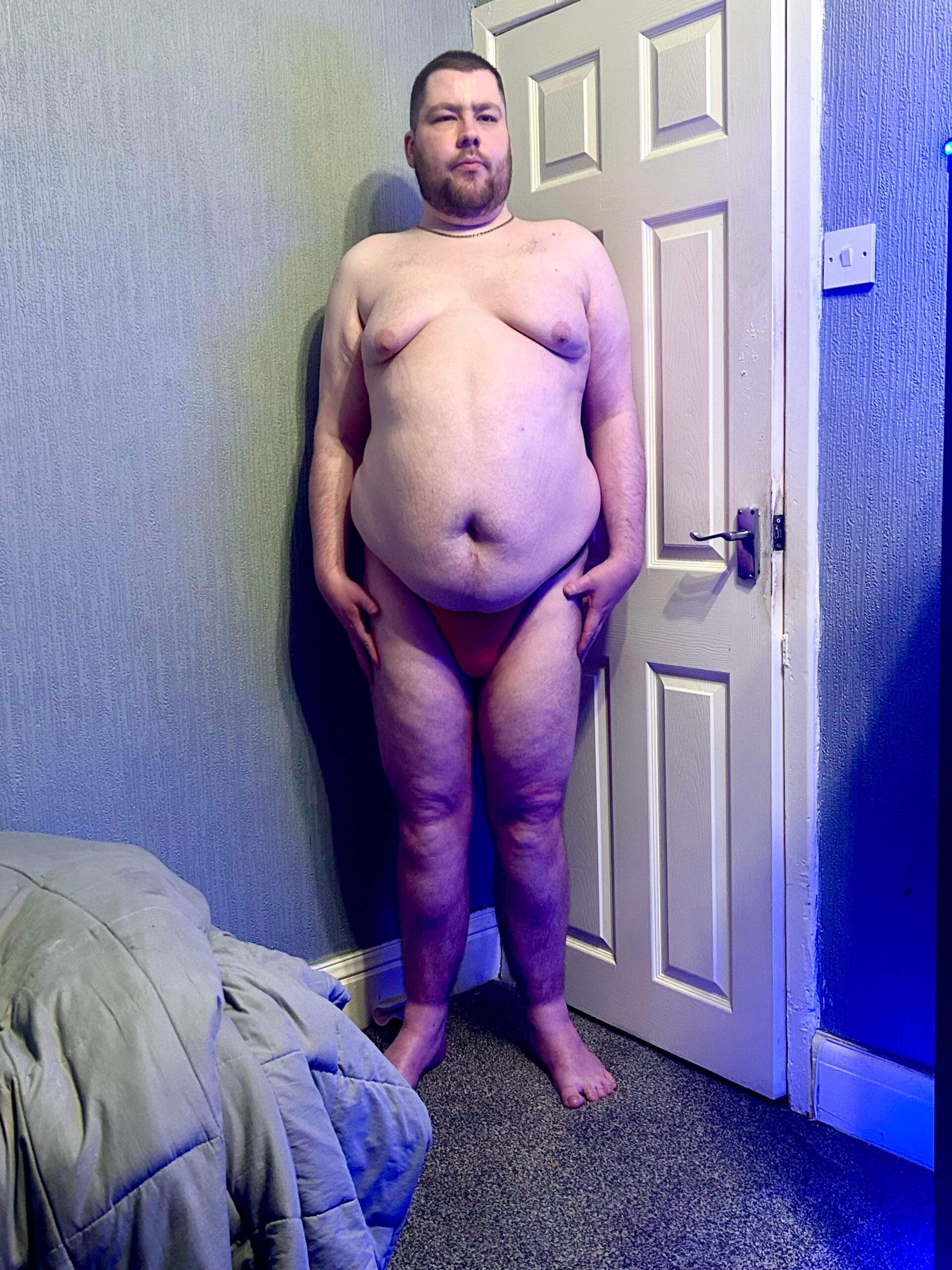 A tall bearded man wearing a bright red Tanga Brief with a black waistband that has Kiniki written in bright white writing on it stood against a white wooden door.