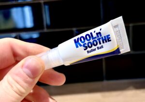 A hand holding a small conical bright white plastic tube that has a Roller Ball at the end of it, Kool ānā Soothe Roller Ball written in bold black writing and a picture of a gradient dark blue and yellow stripe on it, on a white background.