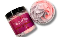 A short cylindrical clear plastic tub full of some gradient bright pink and purple whipped soap with a black plastic lid and a bright red label that has Tales of Toria written in bright white writing, Cherry Slushy Whipped Soap written in bright red writing, and some pictures of some cherries on it, on a white background.