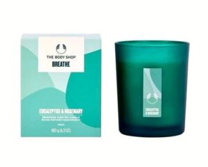 A short cylindrical glass jar full of some bright teal wax with a white label that has The Body Shop Breathe Eucalyptus & Rosemary Renewing Scented Candle written in bold black writing on it next to a medium pale teal cardboard box with a white label that has The Body Shop Breathe Eucalyptus & Rosemary Renewing Scented Candle written in bold black writing on it, on a white background. 