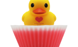A large circular bright white cupcake shaped block of soap that has a bright Pink paper cupcake case around it and a bright Yellow Rubber Duck that has a bright Red Love Heart engraved on it’s chest on top of it, on a white background.
