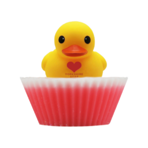 A large circular bright white cupcake shaped block of soap that has a bright Pink paper cupcake case around it and a bright Yellow Rubber Duck that has a bright Red Love Heart engraved on it’s chest on top of it, on a white background.