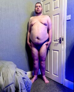 A tall bearded man wearing a pair of bright purple briefs with a bright purple waistband that has Box Menswear written in bold black writing on it stood in front of a white wooden door.