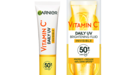 A tall conical bright white plastic tube with a bright yellow plastic lid and a bright yellow label that has Garnier SkinActive Vitamin C Daily Brightening UV Fluid Invisible SPF50+ written in bold black writing on it next to a tall rectangular gradient bright white and yellow cardboard box that has Garnier SkinActive Vitamin C Daily Brightening Fluid Invisible SPF50+ written in bold black writing and some pictures of some slices of some bright yellow Lemons on it, on a white background.