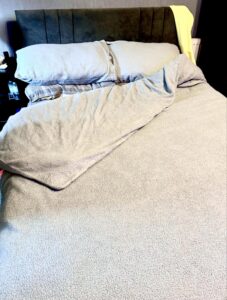 A large rectangular double Bed with a large rectangular plush dark grey headboard and some fluffy dark grey cushions that's covered in a large fluffy dark grey duvet cover, on a white background.
