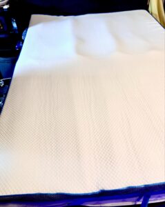 A large rectangular bright white foam mattress topper that has a geometric square and diamond pattern all over it on top of a dark white mattress in front of a large rectangular plush dark grey headboard, on a white background.