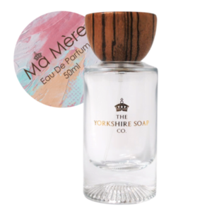 A tall rectangular clear glass bottle full of some clear liquid with a light wooden lid and a white label that has The Yorkshire Soap Company Ma Mere Eau De Parfum written in bold black writing and a picture of a black crown on it, on a white background.