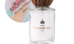 A tall rectangular clear glass bottle full of some clear liquid with a light wooden lid and a white label that has The Yorkshire Soap Company Maman Eau De Parfum written in bold black writing and a picture of a black crown on it, on a white background.