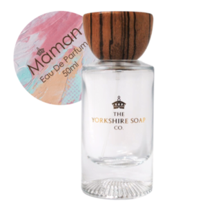 A tall rectangular clear glass bottle full of some clear liquid with a light wooden lid and a white label that has The Yorkshire Soap Company Maman Eau De Parfum written in bold black writing and a picture of a black crown on it, on a white background.