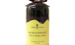 A tall and wide cylindrical plastic bottle full of some dark brown liquid with a silver plastic lid, a dark brown bow tied around it, and a gradient dark brown and bright yellow label that has The Yorkshire Soap Company Melted Chocolate Easter Egg Bath & Shower Creme written in bold black writing and a picture of a black crown on it, on a white background.