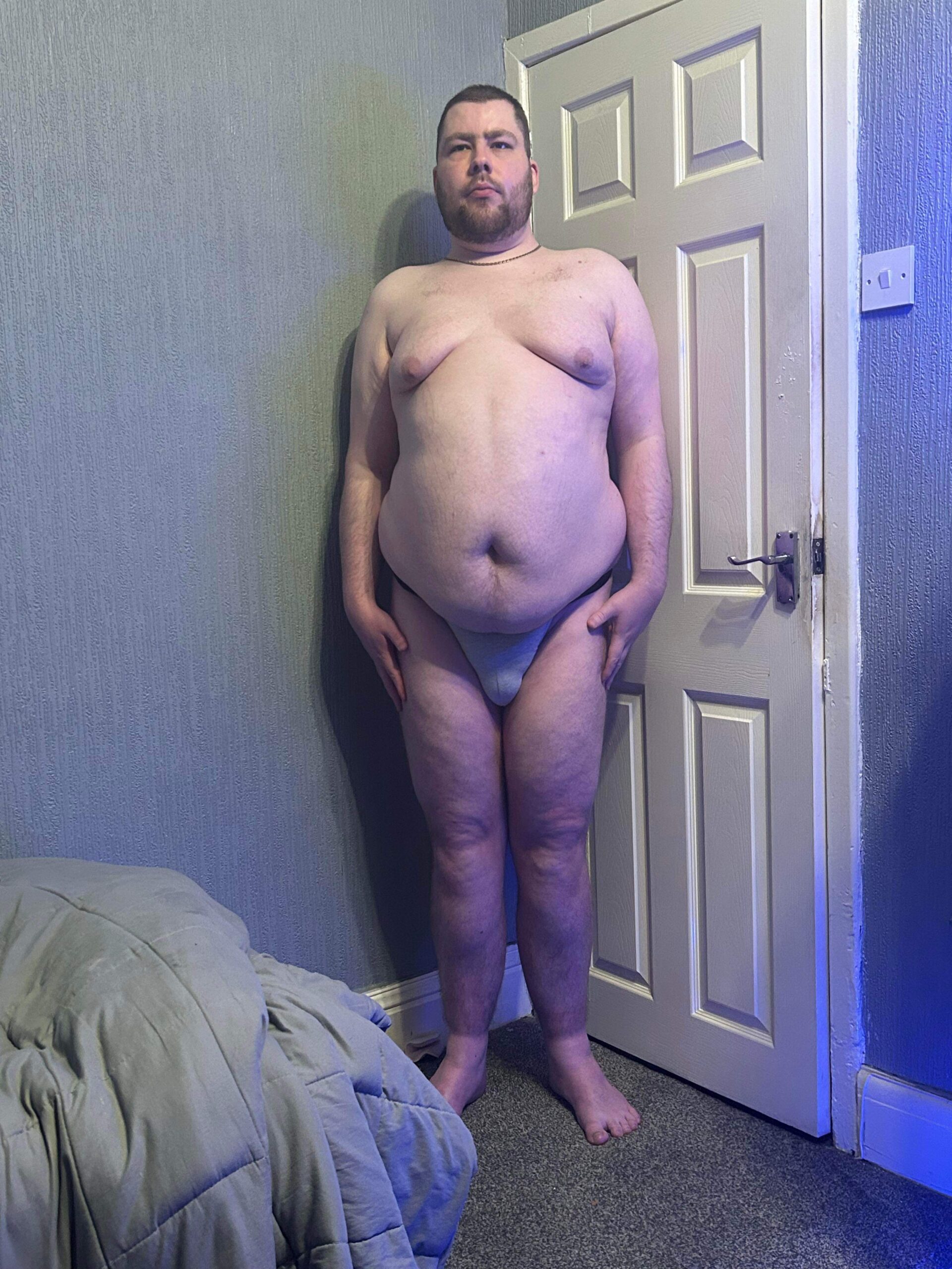 A tall bearded man wearing a bright silver tanga brief with a black waistband that has Kiniki written in bright white writing on it stood against a white wooden door.