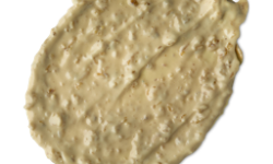 A medium circular puddle of some pale yellow face mask that has some pieces of Porridge running through it, on a white background.