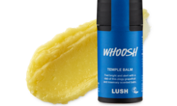A medium circular puddle of some pale yellow temple balm next to a short cylindrical dark blue plastic tube with a black plastic lid that has Lush Whoosh Temple Balm written in bright white writing on it, on a white background.