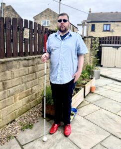 A tall bearded man wearing a pair of silver and black aviator sunglasses with some light green lenses, a light blue short sleeve shirt that has a white diamond print all over it, a dark navy V-neck t-shirt, a pair of dark navy chino trousers and a pair of shiny bright red shoes holding a bright white cane with a red leather handle stood against a dark wooden fence.