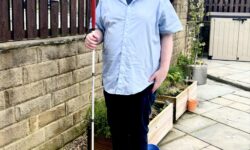 A tall bearded man wearing a pair of silver and black aviator sunglasses with some light green lenses, a light blue short sleeve shirt that has a white diamond print all over it, a dark navy V-neck t-shirt, a pair of dark navy chino trousers and a pair of shiny bright red shoes holding a bright white cane with a red leather handle stood against a dark wooden fence.