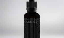 A short cylindrical brown glass bottle with a black rubber pipette lid and a black label that has runesilk elements Metal beard oil written in bold black writing and a picture of a white geometric pattern on it, on a white background.