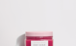 A short square clear plastic tub full of some dark pink body scrub with a bright pink plastic lid and a white label that has Sunday Rain Body Scrub written in bold black writing and Rose written in bright green writing on it, on a white background.