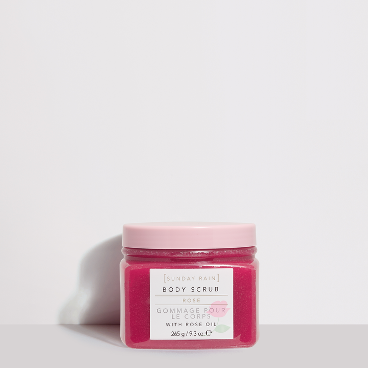 A short square clear plastic tub full of some dark pink body scrub with a bright pink plastic lid and a white label that has Sunday Rain Body Scrub written in bold black writing and Rose written in bright green writing on it, on a white background.