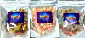 A tall rectangular plastic bag full of some yellow, red and white gummy sweets with a dark purple label that has Sweets written in bright yellow writing and Smiles written in bright pink writing on it next to a tall rectangular plastic bag full of some yellow, orange, pink and green coloured boiled sweets with a dark purple label that has Sweets written in bright yellow writing and Smiles written in bright pink writing on it, and a tall rectangular plastic bag full of some red, orange, yellow and green coloured fizzy sweets with a dark purple label that has Sweets written in bright yellow writing and Smiles written in bright pink writing on it, on a white background.