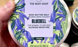 A short cylindrical bright silver plastic tub with a bright silver plastic lid and a bright white label that has The Body Shop Bluebell Body Butter Melt written in dark purple writing and a picture of some dark purple bluebell flowers on it, on a white background.