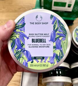 A short cylindrical bright silver plastic tub with a bright silver plastic lid and a bright white label that has The Body Shop Bluebell Body Butter Melt written in dark purple writing and a picture of some dark purple bluebell flowers on it, on a white background.