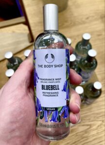 A tall cylindrical clear glass bottle full of some clear liquid with a silver metal lid and a pale purple label that has The Body Shop Bluebell Fragrance Mist written in bold black writing and a picture of some dark purple bluebells on it, on a white background.