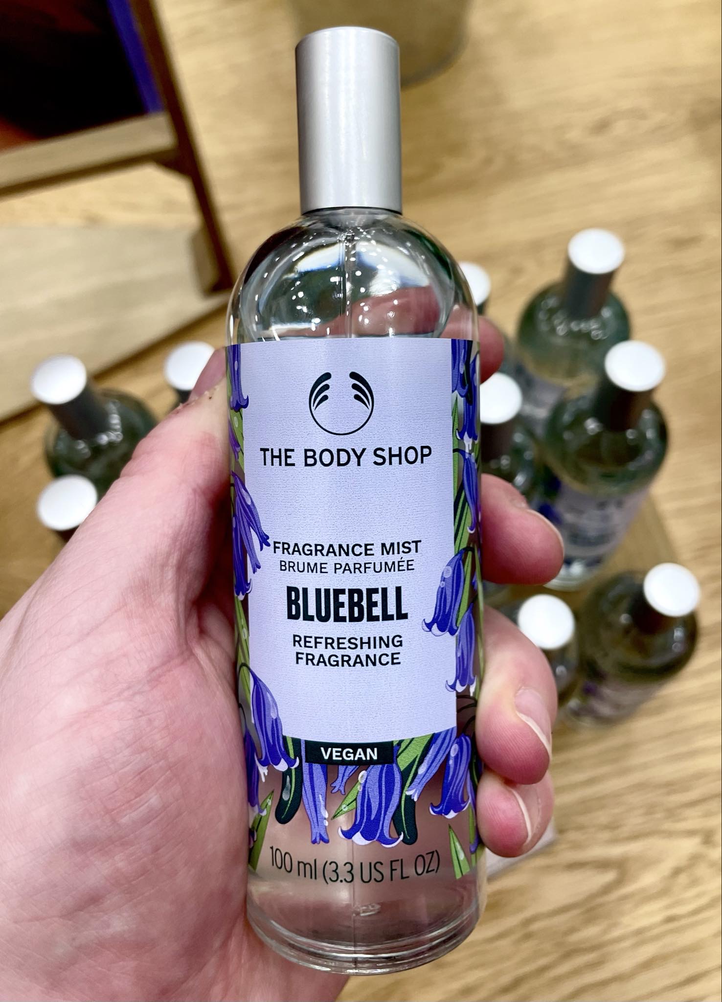 A tall cylindrical clear glass bottle full of some clear liquid with a silver metal lid and a pale purple label that has The Body Shop Bluebell Fragrance Mist written in bold black writing and a picture of some dark purple bluebells on it, on a white background.