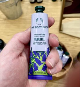 A long and thin conical metallic white tube with a metallic dark green lid and a bright white label that has The Body Shop Bluebell Hand Cream written in dark green writing and some pictures of some dark purple bluebells on it, on a white background.