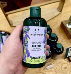 A tall cylindrical dark green plastic bottle with a dark green plastic lid and a bright white label that has The Body Shop written in bright white writing, Bluebell Shower Cream written in bold black writing and some pictures of some dark purple Bluebell Flowers on it, on a white background.