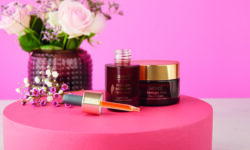 A short cylindrical amber Glass Bottle with a gold Rubber Pipette Lid and a dark brown Label that has Aldi Lacura ProNight Advanced Face Serum written in bright white writing on it next to a short cylindrical amber glass jar with a gold plastic lid and a dark brown label that has Aldi Lacura ProNight+ Face Cream written in bright white writing on it, on a bright pink background.