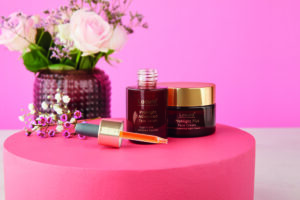 A short cylindrical amber Glass Bottle with a gold Rubber Pipette Lid and a dark brown Label that has Aldi Lacura ProNight Advanced Face Serum written in bright white writing on it next to a short cylindrical amber glass jar with a gold plastic lid and a dark brown label that has Aldi Lacura ProNight+ Face Cream written in bright white writing on it, on a bright pink background.
