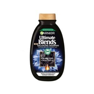 A tall rounded rectangular black plastic Bottle with a gold plastic lid and a bright green label that has Garnier Ultimate Blends Balancing Shampoo with Charcoal & Black Seed Oil written in bright white writing, a picture of a puddle of black charcoal, and a picture of some black seeds on it, on a white background.