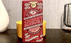 A large rectangular chocolate bar wrapped in a dark red paper wrapper that has The Chocolate Smiths written in bold black writing and Cherry Bakewell written in bright gold writing on it, on a dark wooden cabinet.