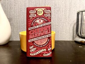 A large rectangular chocolate bar wrapped in a dark red paper wrapper that has The Chocolate Smiths written in bold black writing and Cherry Bakewell written in bright gold writing on it, on a dark wooden cabinet.