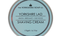 A short cylindrical metallic silver tin with a pastel blue label that has The Yorkshire Soap Company Yorkshire Lad Shaving Cream written in bright white writing on it, on a white background.