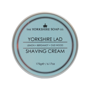 A short cylindrical metallic silver tin with a pastel blue label that has The Yorkshire Soap Company Yorkshire Lad Shaving Cream written in bright white writing on it, on a white background.